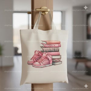 May include: Cream-colored tote bag featuring a graphic of pink high-top sneakers and a stack of books. The books have the words "STANDING ON BUSINESS" printed on them. The bag has a simple design with long handles.