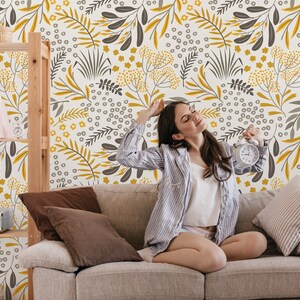 Puede incluir: Una mujer se sienta en un sof&aacute; gris claro frente a una pared con un patr&oacute;n floral. El patr&oacute;n presenta hojas y flores amarillas y grises sobre un fondo blanco. La mujer lleva una camisa a rayas blancas y grises y tiene los ojos cerrados.