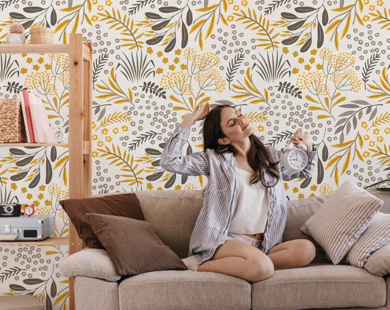 Puede incluir: Una mujer se sienta en un sof&aacute; gris claro frente a una pared con un patr&oacute;n floral. El patr&oacute;n presenta hojas y flores amarillas y grises sobre un fondo blanco. La mujer lleva una camisa a rayas blancas y grises y tiene los ojos cerrados.