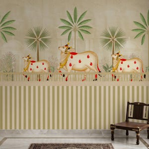 May include: A decorative wall mural featuring three stylized white cows with red handprints, adorned with gold and red accents. Palm trees and a white fence are in the background, with a striped pattern below. A wooden chair is in the foreground.