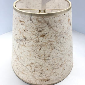 May include: A beige lampshade with a textured, paper-like surface. The shade has a gold trim around the top and bottom.