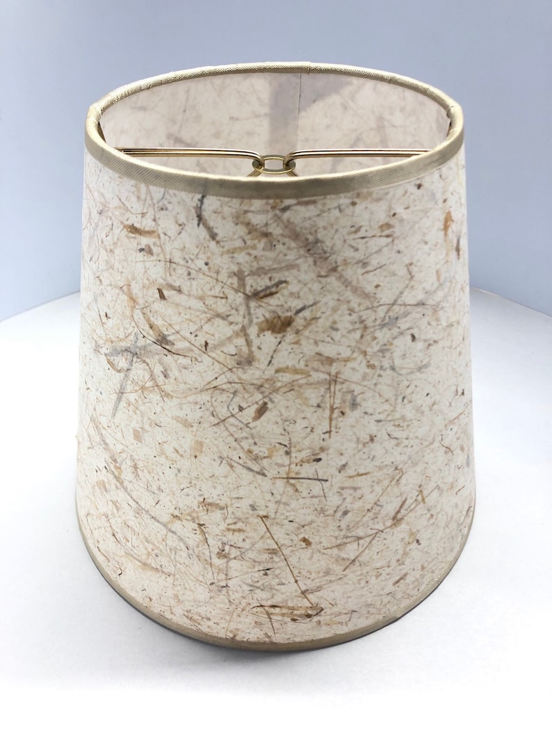 May include: A beige lampshade with a textured, paper-like surface. The shade has a gold trim around the top and bottom.