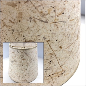May include: A beige lampshade with a textured surface made of natural fibers. The shade has a slightly rounded shape and a gold trim around the top edge.