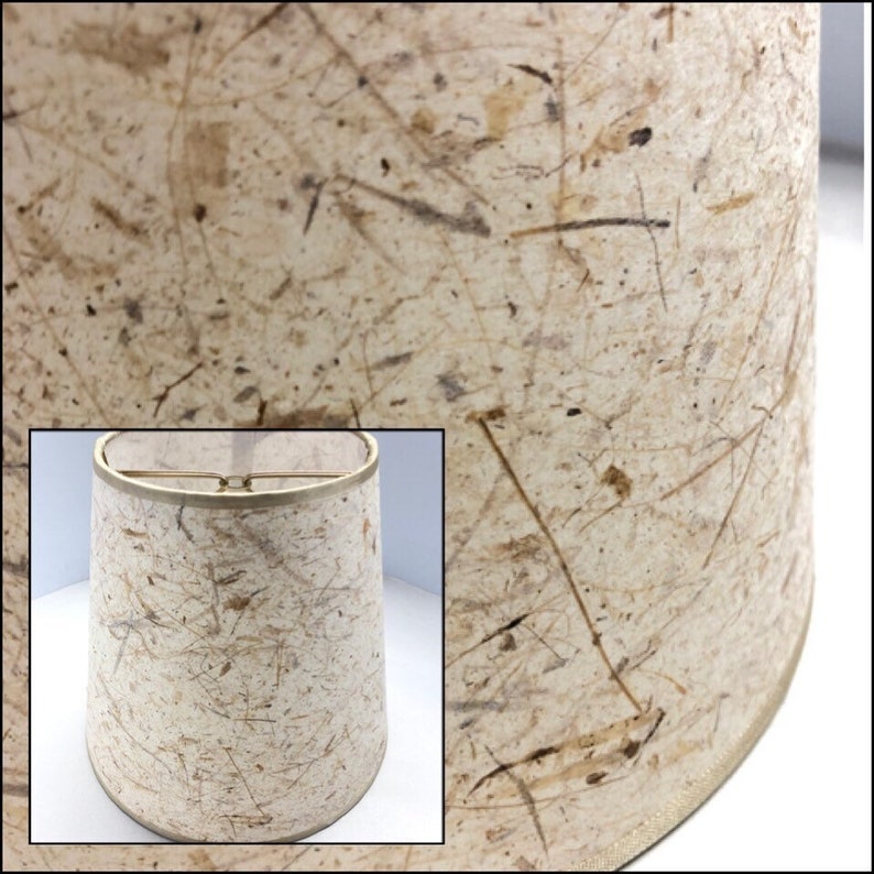 May include: A beige lampshade with a textured surface made of natural fibers. The shade has a slightly rounded shape and a gold trim around the top edge.