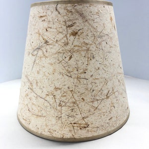 May include: A beige lampshade with a textured surface made of natural fibers. The shade has a slightly curved bottom edge.
