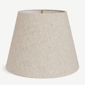 May include: A beige lampshade with a tapered design. The lampshade is made of a textured fabric, and the interior is white. The lampshade has a brass-colored fitting at the top.