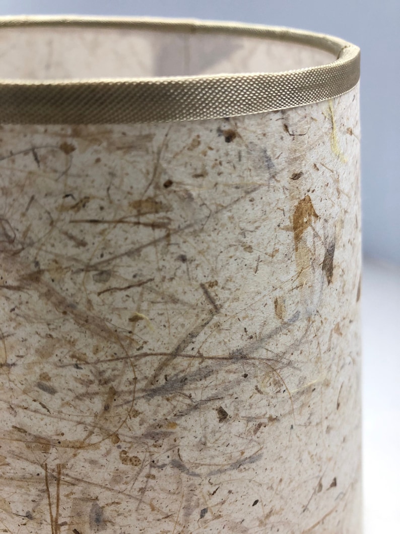 May include: A beige lampshade with a textured surface made of natural fibers. The lampshade has a brown trim around the top edge.