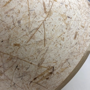 May include: A cream-colored lampshade with a textured surface made of natural fibers. The shade has a gold-colored trim around the edge.