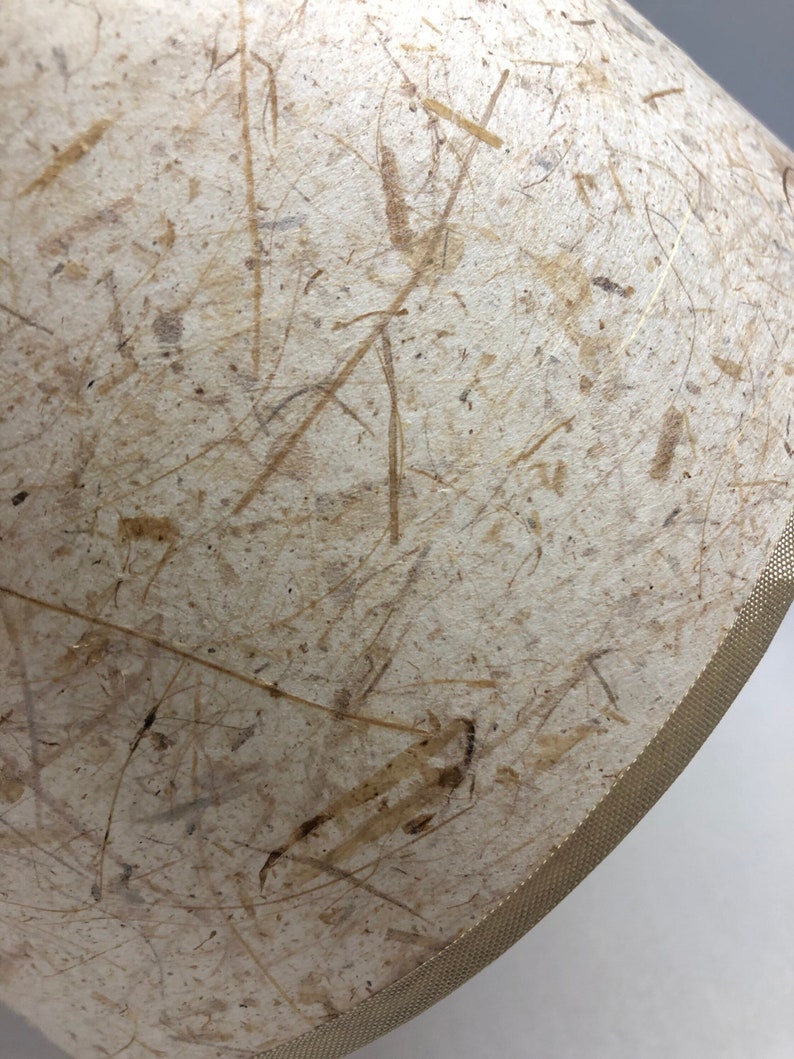 May include: A cream-colored lampshade with a textured surface made of natural fibers. The shade has a gold-colored trim around the edge.