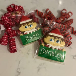 May include: Two elf-shaped cookies, each holding a green sign with a name. One says "Badin" and the other "Bentley." The cookies have red hats and brown hair. Red and white ribbon bows are in the background.