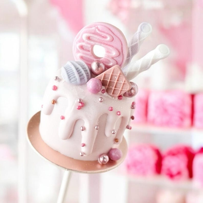 Pu&ograve; includere: Un cake pop rosa con glassa bianca e codette rosa. Il cake pop &egrave; sormontato da una ciambella rosa, un biscotto waffle, un vortice rosa e bianco e una caramella viola. Il cake pop &egrave; su un supporto dorato.