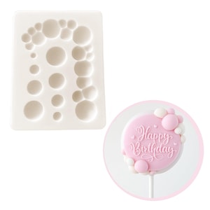 May include: White silicone mold with multiple round cavities of varying sizes. The mold is designed to create bubble-shaped decorations for cakes or other desserts. A pink cake pop with white and pink bubble decorations is shown next to the mold. The cake pop is decorated with the words "Happy Birthday".