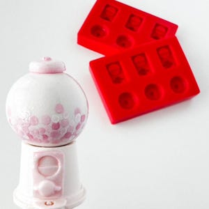 May include: A white and pink gumball machine with a round top and a pink base. The gumball machine has a pink and white polka dot design. Two red silicone molds are in the background.
