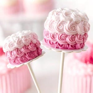 Pu&ograve; includere: Due cake pops glassati rosa e bianchi con un design a spirale. I cake pops sono su bastoncini bianchi e hanno una base di carta bianca.