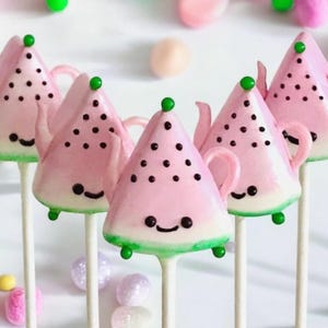 K&ouml;nnte beinhalten: Vier rosafarbene Wassermelonen-Cake-Pops mit gr&uuml;nen Bl&auml;ttern und schwarzen Samen. Jeder Cake-Pop ist in Form einer Teekanne mit einem l&auml;chelnden Gesicht geformt und befindet sich auf einem wei&szlig;en Stiel.