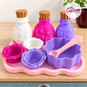May include: A set of three small bottles with brown caps, three flower-shaped bowls, and a light pink spoon on a pink tray. The white bottle has a daisy, the pink bottle has lily of the valley, and the purple bottle has a butterfly design. KM Custom Designs logo.