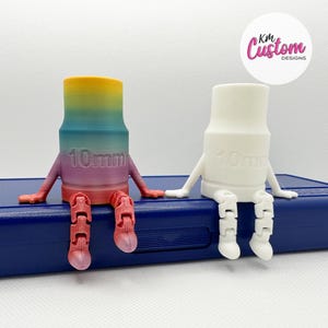 May include: Two 3D printed figurines, one white and one with a rainbow gradient, both with articulated legs and arms. The figurines are sitting on a blue surface and both have "10mm" printed on their bodies.