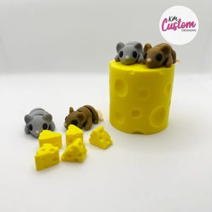 May include: A yellow cheese block with two mice on top and five small cheese wedges on the side. The cheese block has a pattern of holes. The mice are grey and brown. The text "Kim Custom Designs" is in the top right corner of the image.
