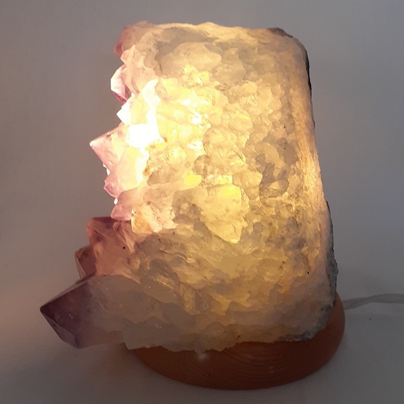 May include: A large, clear amethyst crystal lamp with a wooden base. The crystal is illuminated from within, casting a warm glow. The amethyst has a purple hue and is naturally formed.