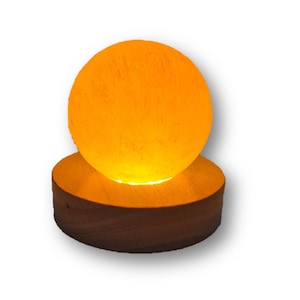 Könnte beinhalten: Eine runde, orangefarbene Salzlampe mit einem Holzsockel. Die Lampe ist eingeschaltet und leuchtet.