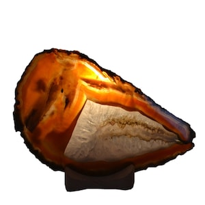 Könnte beinhalten: Eine natürliche Agat-Geodenlampe mit einem warmen orange-weißen Farbschema. Die Geode wird von innen beleuchtet und erzeugt so ein sanftes Leuchten. Die Geode ist auf einem Holzsockel montiert.