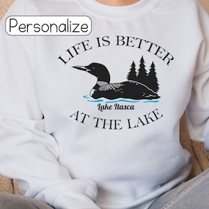 May include: White t-shirt with black text that reads "Life is better at the lake" with a black loon illustration and the words "Lake Itasca" underneath.