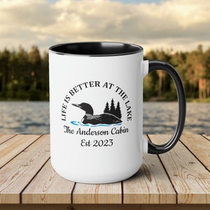 May include: White ceramic mug with a black rim and handle. The mug features a black and white illustration of a loon swimming in a lake with the text "Life is better at the lake" and "The Anderson Cabin Est 2023".