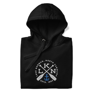 Puede incluir: Sudadera con capucha negra con un logotipo bordado en blanco. El logotipo es un c&iacute;rculo con el texto "Lake Norman, North Carolina" y un dise&ntilde;o de palas cruzadas con un ancla en el centro. El texto "ESTD. 1963" est&aacute; debajo del ancla.