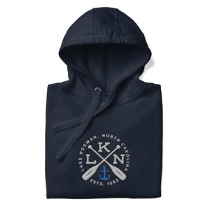 Puede incluir: Sudadera con capucha azul marino con un logotipo bordado en blanco. El logotipo presenta un c&iacute;rculo con el texto "Lake Norman, North Carolina" y un dise&ntilde;o de remos cruzados con un ancla en el centro. El texto "ESTD. 1963" est&aacute; debajo del ancla.