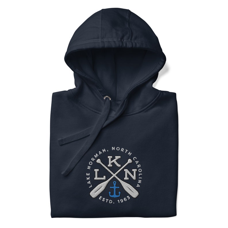 Puede incluir: Sudadera con capucha azul marino con un logotipo bordado en blanco. El logotipo presenta un c&iacute;rculo con el texto "Lake Norman, North Carolina" y un dise&ntilde;o de remos cruzados con un ancla en el centro. El texto "ESTD. 1963" est&aacute; debajo del ancla.