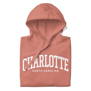 May include: A coral pink hoodie with a white print that says "CHARLOTTE NORTH CAROLINA" in a collegiate font.