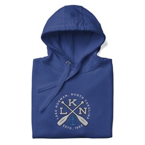 Puede incluir: Sudadera con capucha azul real con un logotipo circular blanco que presenta el texto "Lake Norman, North Carolina" y un dise&ntilde;o de remos cruzados con un ancla en el centro. El texto "ESTD. 1965" est&aacute; debajo del dise&ntilde;o.