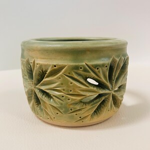 May include: A cylindrical ceramic vessel featuring a light green glaze with subtle yellow undertones. The exterior surface is decorated with carved, star-shaped floral patterns and small punctured dots, creating open cutouts in the clay body.