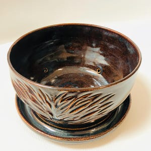 May include: A handcrafted, dark brown ceramic planter with a matching saucer. The planter features a carved leaf design around the rim and three drainage holes. The glazed finish gives it a glossy appearance.