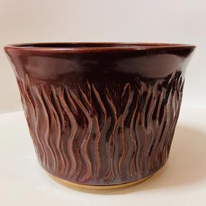 May include: A dark burgundy ceramic planter with a glossy finish. The exterior features a textured, vertical wave pattern. The planter has a wide opening and a slightly flared shape, suitable for various plants.