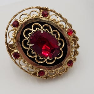 May include: A gold-toned brooch with a red rhinestone in the center, surrounded by a filigree design and smaller red rhinestones.