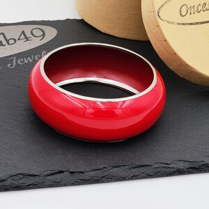 May include: A vibrant red bangle bracelet with a silver-colored interior and trim. The bracelet is glossy and sits on a black surface. A round, tan box is in the background.
