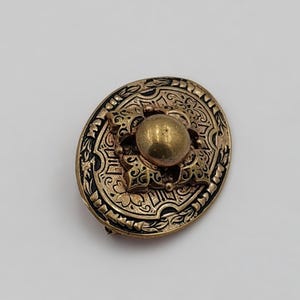 May include: An antique gold-toned brooch with an oval shape. The brooch features intricate floral and scrollwork designs around the edges and a central raised sphere. The metal has a tarnished appearance, suggesting age and wear. A classic piece of jewelry.