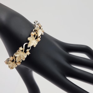 May include: Gold-toned bracelet featuring a leaf design with small, round, peach-colored beads. The bracelet is displayed on a black hand, showcasing its intricate details and elegant style. The clasp is adorned with clear crystals.