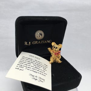 May include: A gold-colored teddy bear pin with a red bow, displayed in a black velvet box. The box has the text "R.J. Graziano" and a logo. A small card with text is visible.