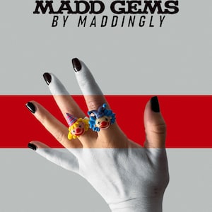 Könnte beinhalten: Zwei Ringe mit Clown-Motiv, mit lächelnden Clown-Gesichtern mit Partyhüten und lockigem Haar, werden auf einer Hand mit schwarzem Nagellack gezeigt. Die Ringe haben gelbe, blaue und rote Akzente. Im Hintergrund steht der Text "MADD GEMS BY MADDINGLY".