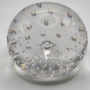May include: Clear glass paperweight with embedded bubbles and a central teardrop-shaped feature. The spherical paperweight has a smooth, rounded shape and is filled with numerous small bubbles, creating a unique visual effect. The paperweight is a decorative item.