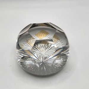May include: Clear, faceted glass paperweight with a rounded shape. The paperweight features a repeating pattern of starburst designs and diamond-shaped facets. The interior has a gold-colored accent, adding a touch of elegance to the design.