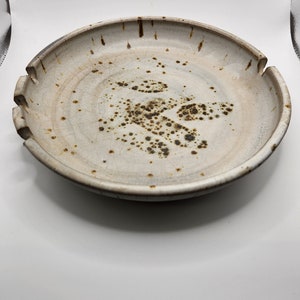 May include: A handmade ceramic plate with a wide, slightly irregular rim. The plate is off-white with brown speckles and a darker brown rim. The design includes cut-out sections on the rim, adding a unique touch to this rustic, handcrafted piece.