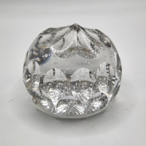 May include: Clear glass paperweight with a rounded shape and a textured, cut-glass design. The paperweight has a series of circular indentations and internal bubbles, creating a decorative effect. The glass is transparent, allowing light to pass through.