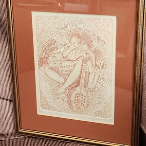 May include: Framed artwork featuring two cherubic figures with wings, one holding a violin. The artwork is in sepia tones, with intricate line work and the words "HIVER ONTES" visible. The frame is gold-colored.