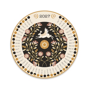 May include: A circular 2027 calendar featuring a central illustration of a winged rabbit surrounded by floral motifs and a sun icon. The outer border displays lunar phases and dates in black text against a cream background, with a black inner circle.