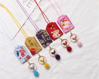May include: Five colorful Chinese New Year charm necklaces with different designs and colors. Each necklace features a small, round charm with a furry pom-pom.
