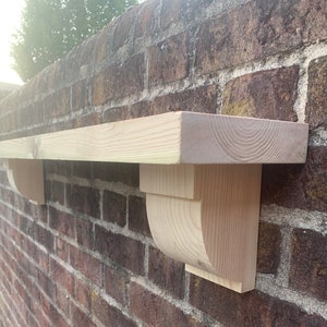 May include: A wooden shelf, approximately 100 cm long, fixed to a brick wall. The shelf is supported by two decorative brackets. The shelf is made of untreated wood. The brackets have a curved design.