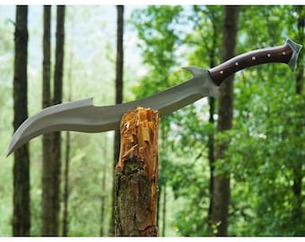 Può includere: Una spada color argento con una lama curva e un'impugnatura in legno marrone. La spada è conficcata in un tronco d'albero in un ambiente boschivo.